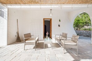Une terrasse couverte, meublée de meubles en osier, avec une table ornée d’un vase et une vue dégagée à travers une arche.