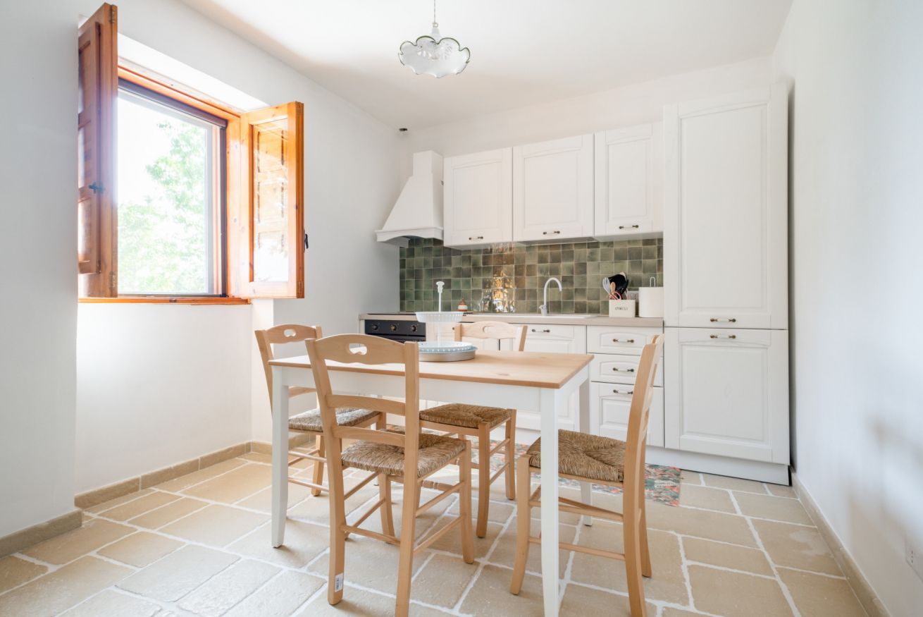 A kitchen with white cabinets, a window with wooden shutters, a dining table with chairs, and a tiled floor.