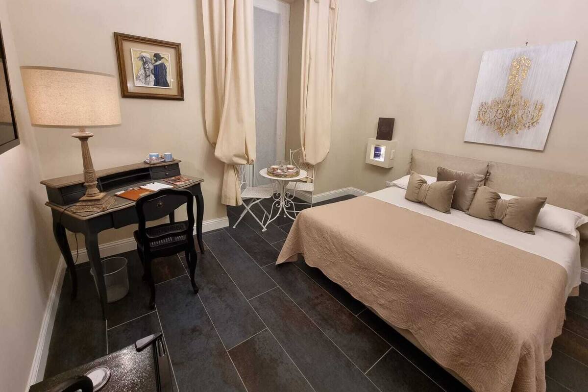 Habitación de hotel con cama, escritorio con lámpara, silla, mesita con bandeja y un cuadro en la pared.