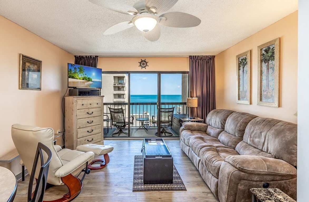 Un salon comprenant un canapé, un fauteuil, un téléviseur, un ventilateur de plafond et une vue sur l’océan grâce à de grandes fenêtres.