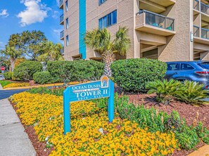A sign for Ocean Dunes Tower II, a multi-story building with balconies, surrounded by a landscaped garden with yellow flowers.