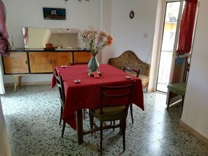 A dining area with a table covered in a red tablecloth, wooden chairs, a vase with flowers, and a patterned sofa.