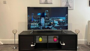 A television on a black stand with decorative jars, set against a wall with framed cityscape pictures. - Jacuzzi Spa! Stunning Entire Guest House - Netflix (Derby)