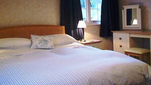A neatly arranged bedroom with a large bed, a bedside table with a lamp, a dresser, and a window with curtains.