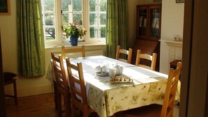 A dining room with a table set for four, wooden chairs, a window with curtains, and a cabinet with books.