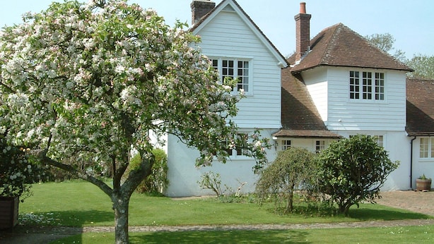 A two-story white house with a brown roof, surrounded by a well-maintained lawn and flowering trees.