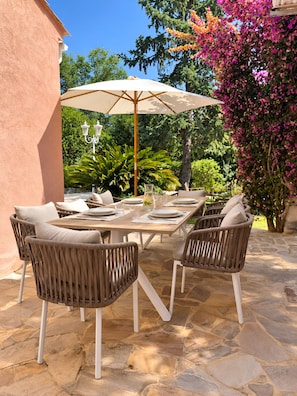 Een terras met een stenen vloer, rieten stoelen, een houten tafel voor vier personen en een grote parasol die schaduw geeft.