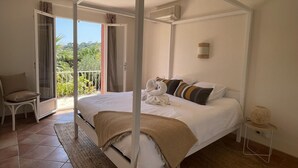 Un dormitorio con cama con dosel, un balcón con vistas a la vegetación y una pequeña mesa con lámpara.