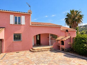Une villa rose avec un toit recouvert de tuiles, une entrée pavée de pierres et un palmier en arrière-plan.