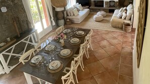 Un comedor amplio con una larga mesa dispuesta para seis personas, rodeada de sillas blancas. Se observa una pared de piedra y una chimenea al fondo.