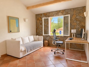 Ein Zimmer mit einem weißen Sofa, einem Schreibtisch mit Computer, einer Steinwand und einem Fenster mit Blick auf Grünflächen.