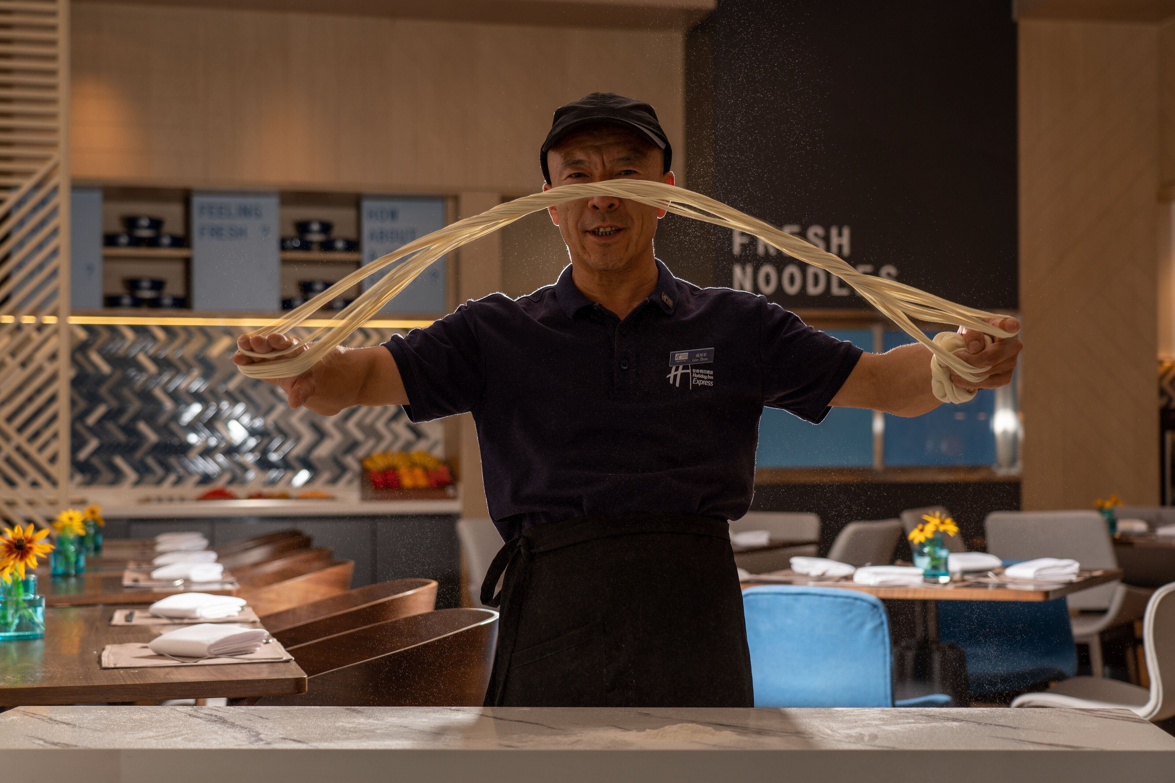 A person demonstrating the preparation of a noodle dish in a kitchen setting with a modern design, including a patterned backsplash and a counter with a bowl of noodles.