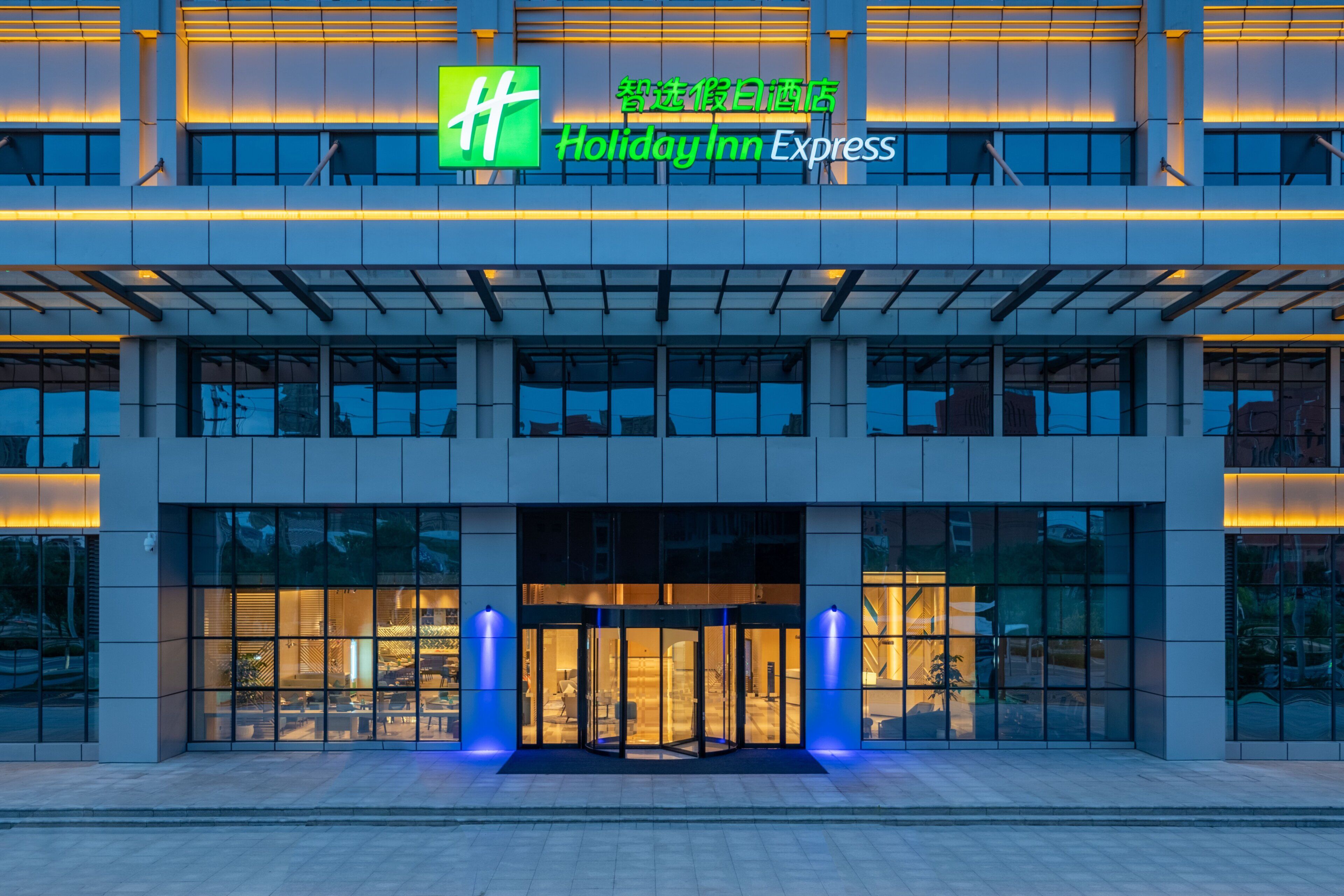 Um hotel Holiday Inn Express com fachada moderna, grandes janelas de vidro e entrada destacada com caminho iluminado em azul.