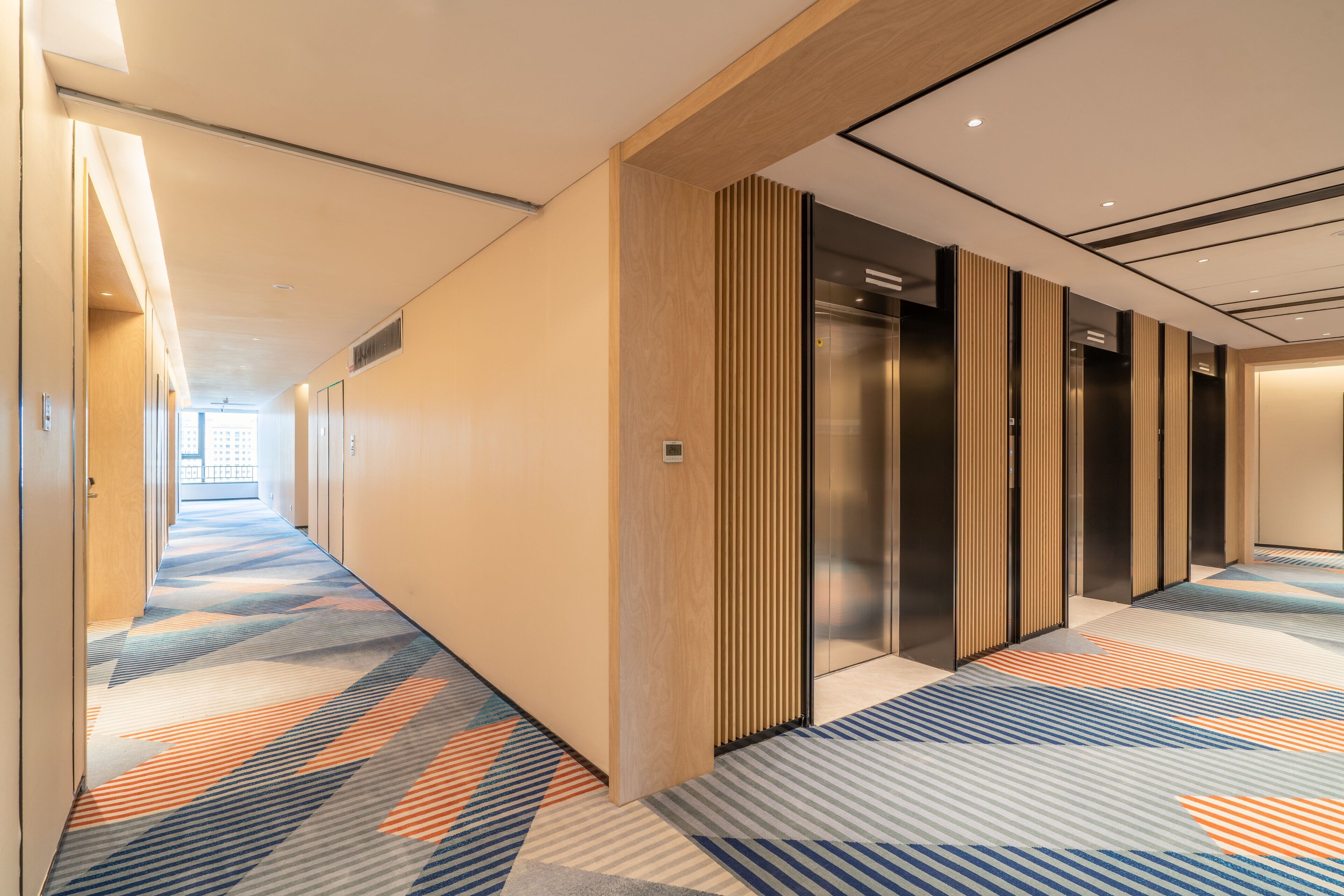 Un couloir d’hôtel avec un tapis à motifs, des murs recouverts de panneaux en bois et un ascenseur en verre.
