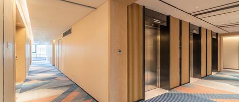 Un couloir d’hôtel avec un tapis à motifs, des murs recouverts de panneaux en bois et un ascenseur en verre.