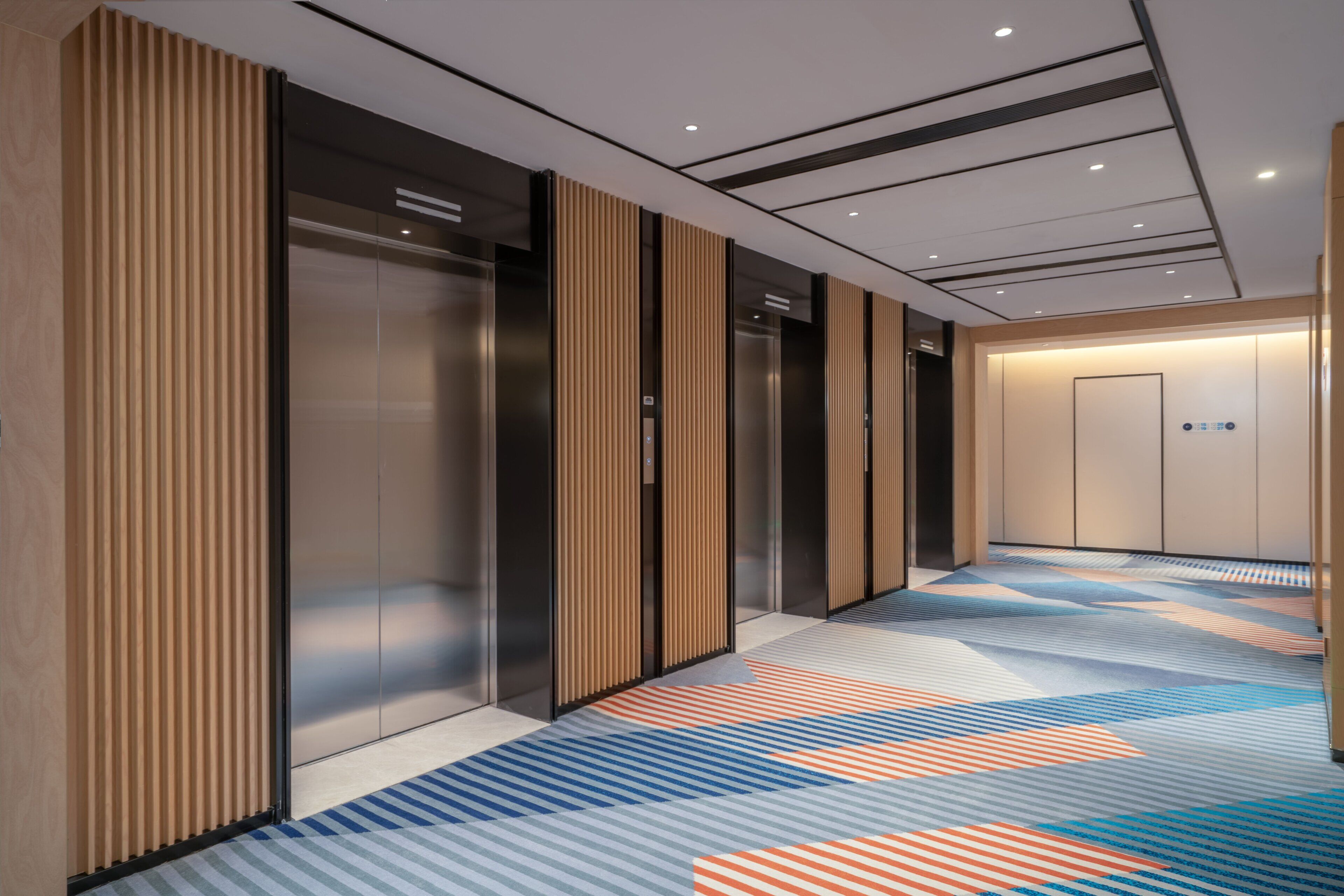 A modern elevator area with metallic elevator doors, a patterned carpet, and a plain wall with a door at the end.