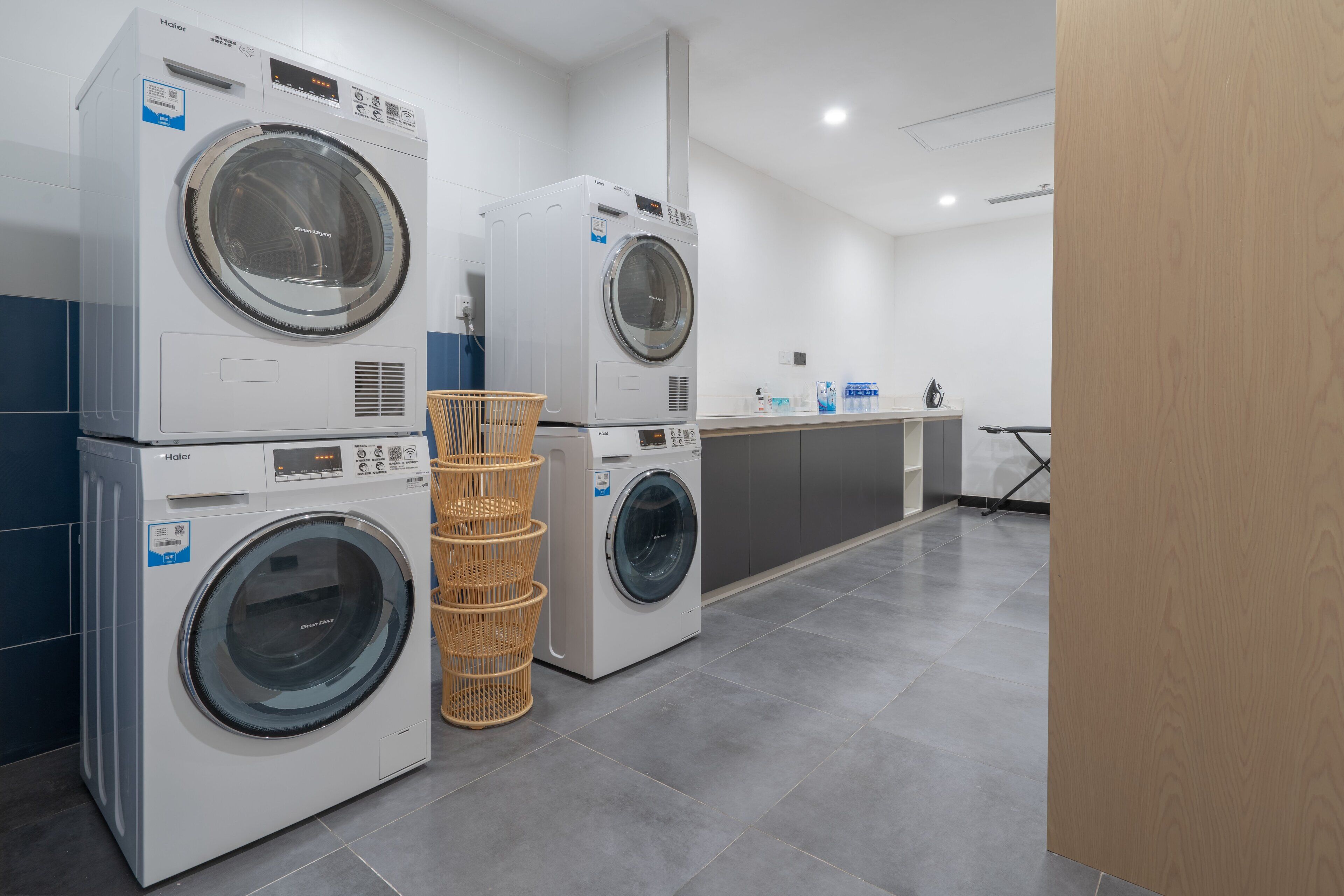 A laundry area with two stacked front-loading washers and dryers, a wicker laundry basket, and a countertop with various items.