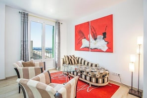 A living room with a sofa, chairs, a glass coffee table, and a red rug. There are two large windows with curtains, a painting on the wall, and a floor lamp.