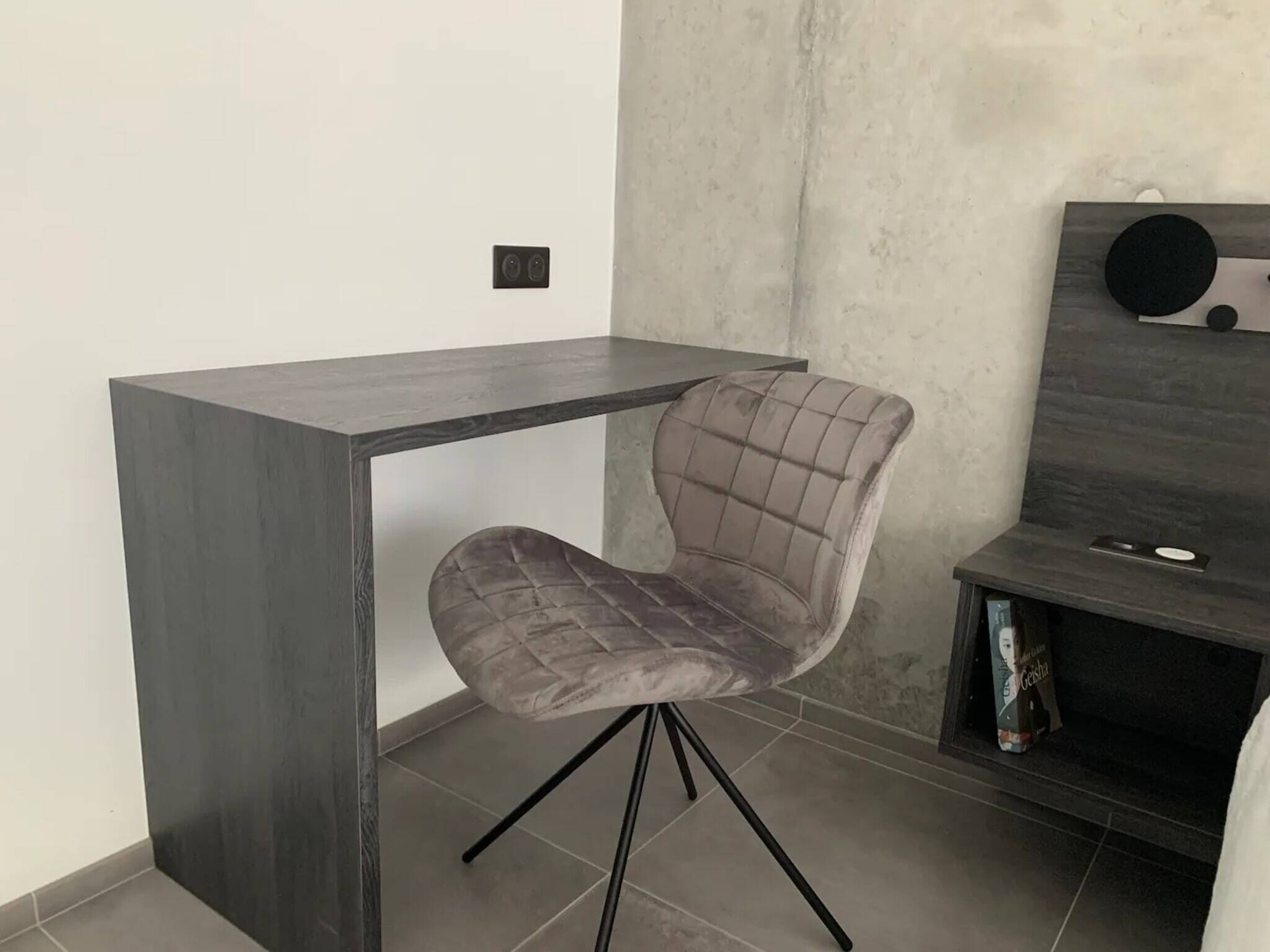 A modern room with a grey upholstered chair, a dark wooden desk, and a bookshelf with books and a speaker.