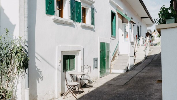 A narrow street with white buildings, green shutters, and a small table with chairs outside.