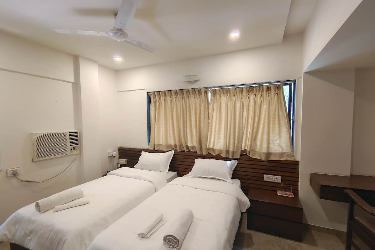A hotel room with two beds, a ceiling fan, an air conditioning unit, a window with curtains, and a bedside table.