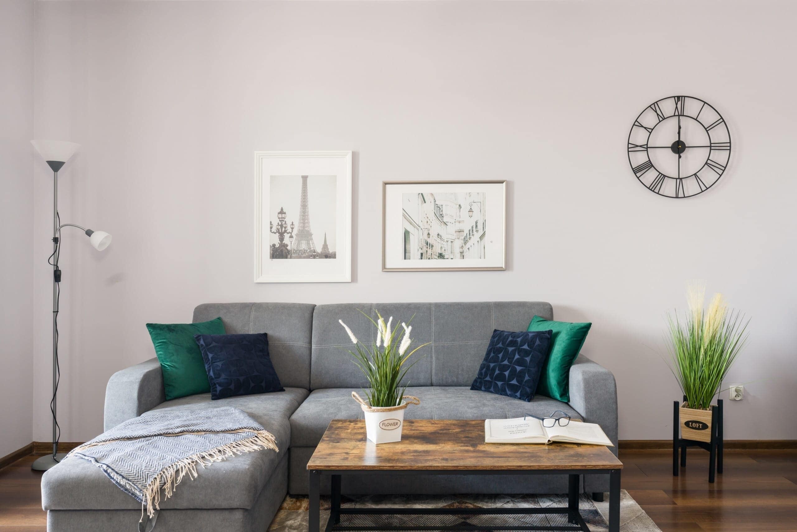 A living room with a grey sofa, two framed pictures, a wooden coffee table with a book and glasses, and a wall clock.