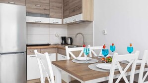 Moderni keittiö, jossa on valkoiset kaapit, ruostumattomasta teräksestä valmistettu jääkaappi ja puinen ruokapöytä valkoisine tuoleineen.