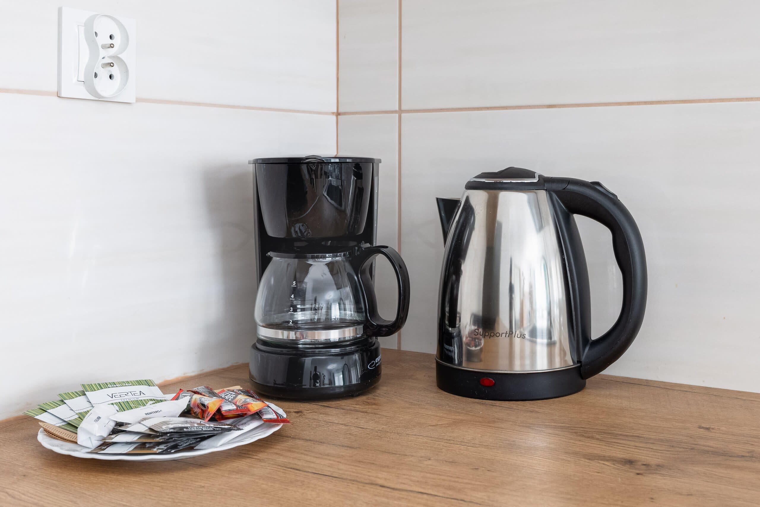 A coffee maker, a kettle, and a plate with coffee packets on a wooden table.