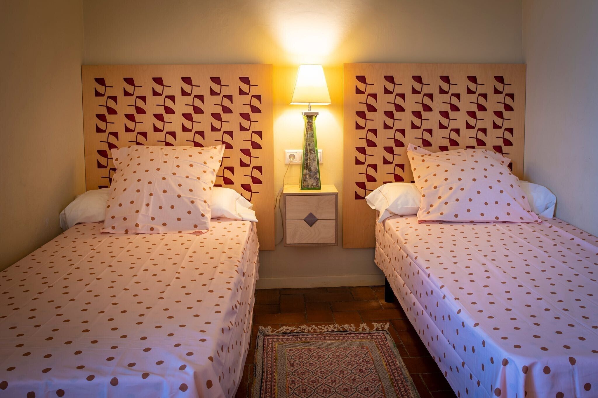 Une chambre d’hôtel avec deux lits, chacun recouvert d’une couvre-lit à pois, une table de chevet avec une lampe et un tapis à motifs.