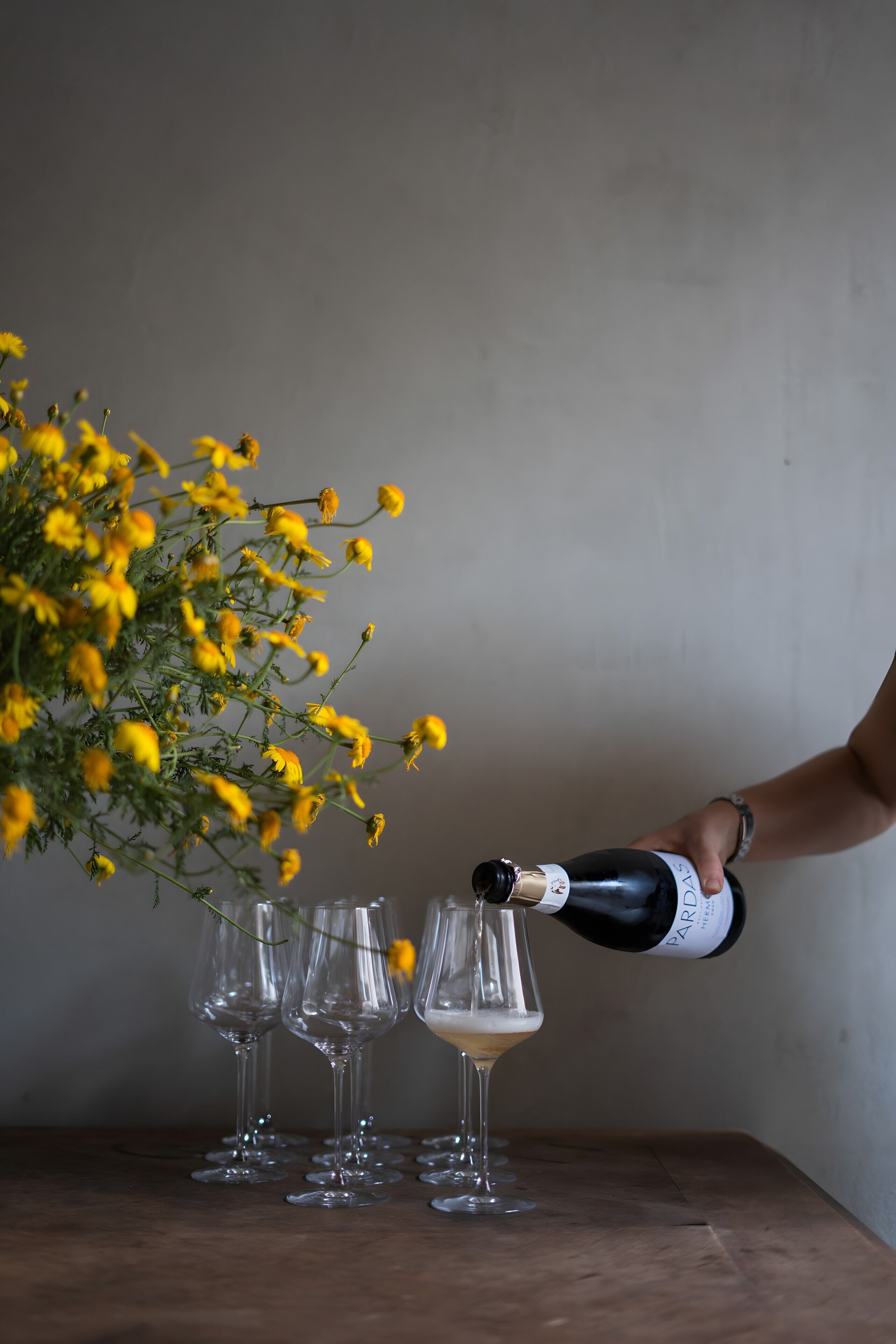 A hand pouring champagne into a glass, with a bouquet of yellow flowers beside it.
