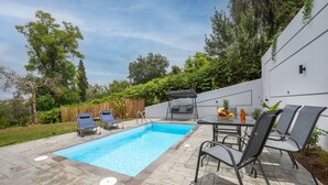 A rectangular swimming pool with a tiled deck, surrounded by lounge chairs and a table with chairs.