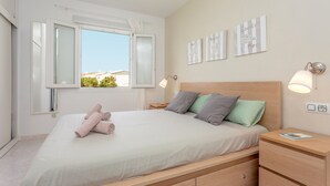 A bedroom with a bed, pillows, a nightstand, a bedside table, a window with curtains, and wall art. - Apartment "Los Delfines Biniforcat" with Private Covered Terrace and Shared Pool (Ciudadela)