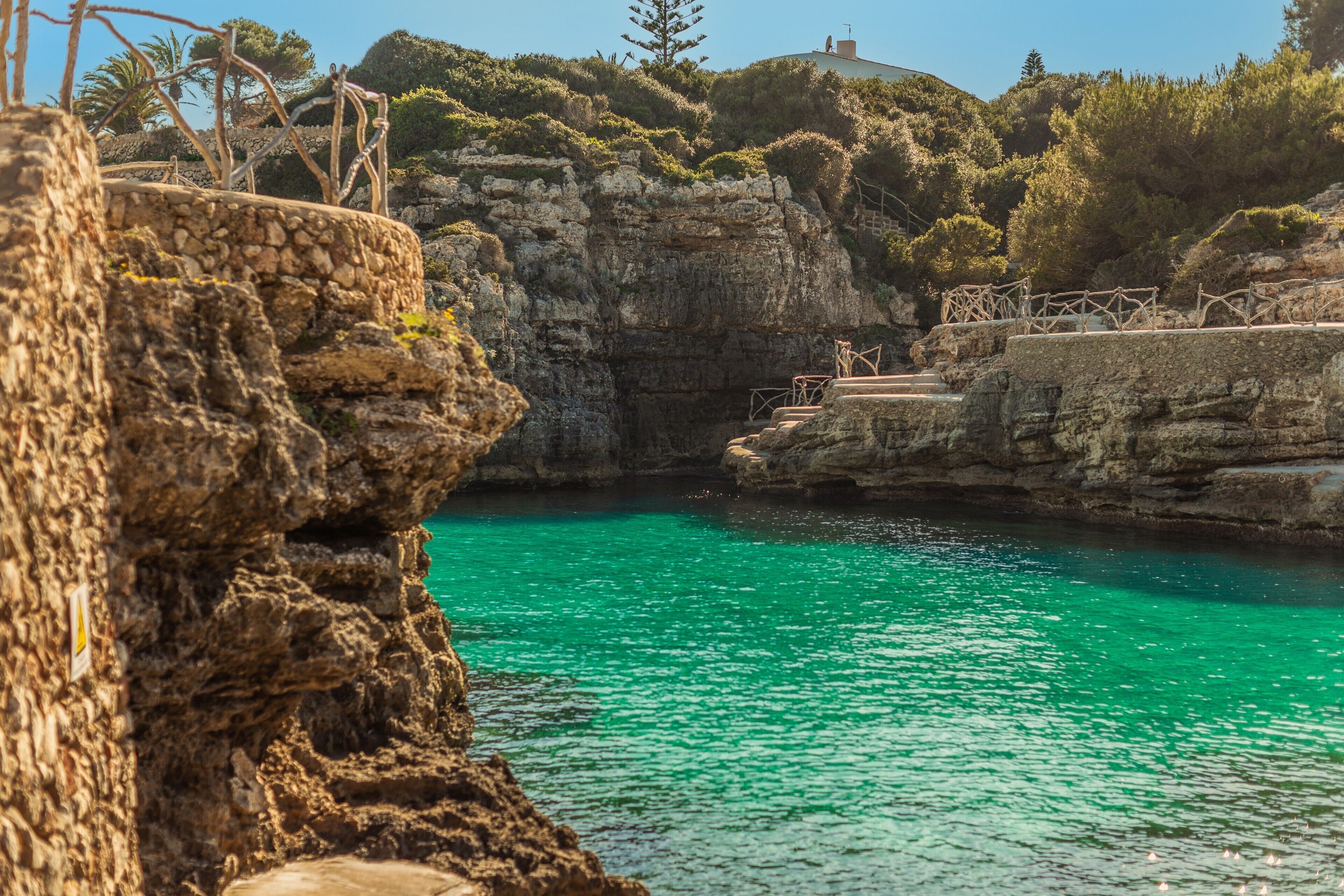 Vista costera con un mar turquesa transparente, acantilados rocosos y una escalera de piedra que desciende hacia el agua.