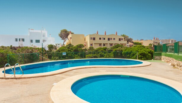 A swimming pool with a curved design, surrounded by a fence and greenery, with buildings in the background. - Apartment "Los Delfines Biniforcat" with Private Covered Terrace and Shared Pool (Ciudadela)