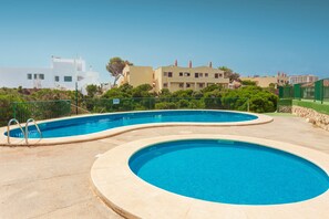 Une piscine au design incurvé, entourée d’une clôture et de verdure, avec des bâtiments en arrière-plan.