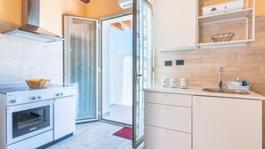Una cucina moderna con forno integrato, una porta vetrata che dà su un balcone e un piano di lavoro bianco con lavello e scaffali contenitori.