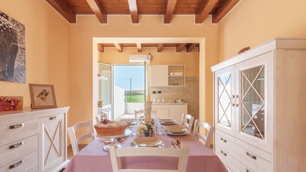 Zona pranzo con tavolo apparecchiato per quattro, cucina sullo sfondo e soffitto in legno.