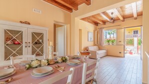Zona pranzo con tavolo imbandito per quattro persone, sedie bianche, soffitto in legno e vista su un balcone con piante.
