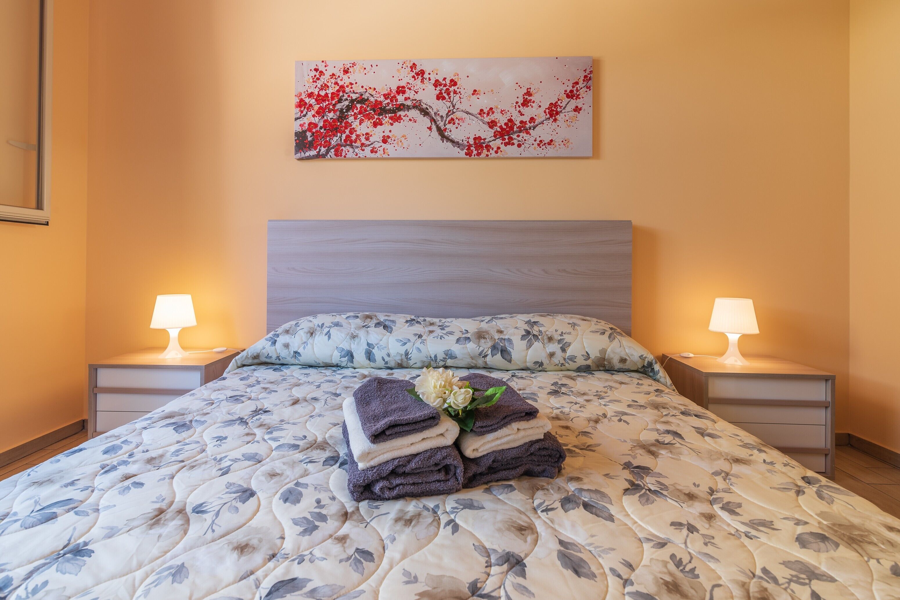 A neatly made bed with floral bedding, two bedside lamps, a wall-mounted painting, and a small bedside table with a lamp.
