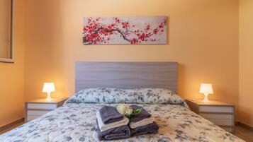 A neatly made bed with floral bedding, two bedside lamps, a wall-mounted painting, and a small bedside table with a lamp.