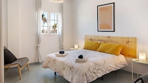 Een slaapkamer met een bed, een stoel, een nachtkastje, een lamp, een raam met gordijnen en een aan de muur hangend schilderij.