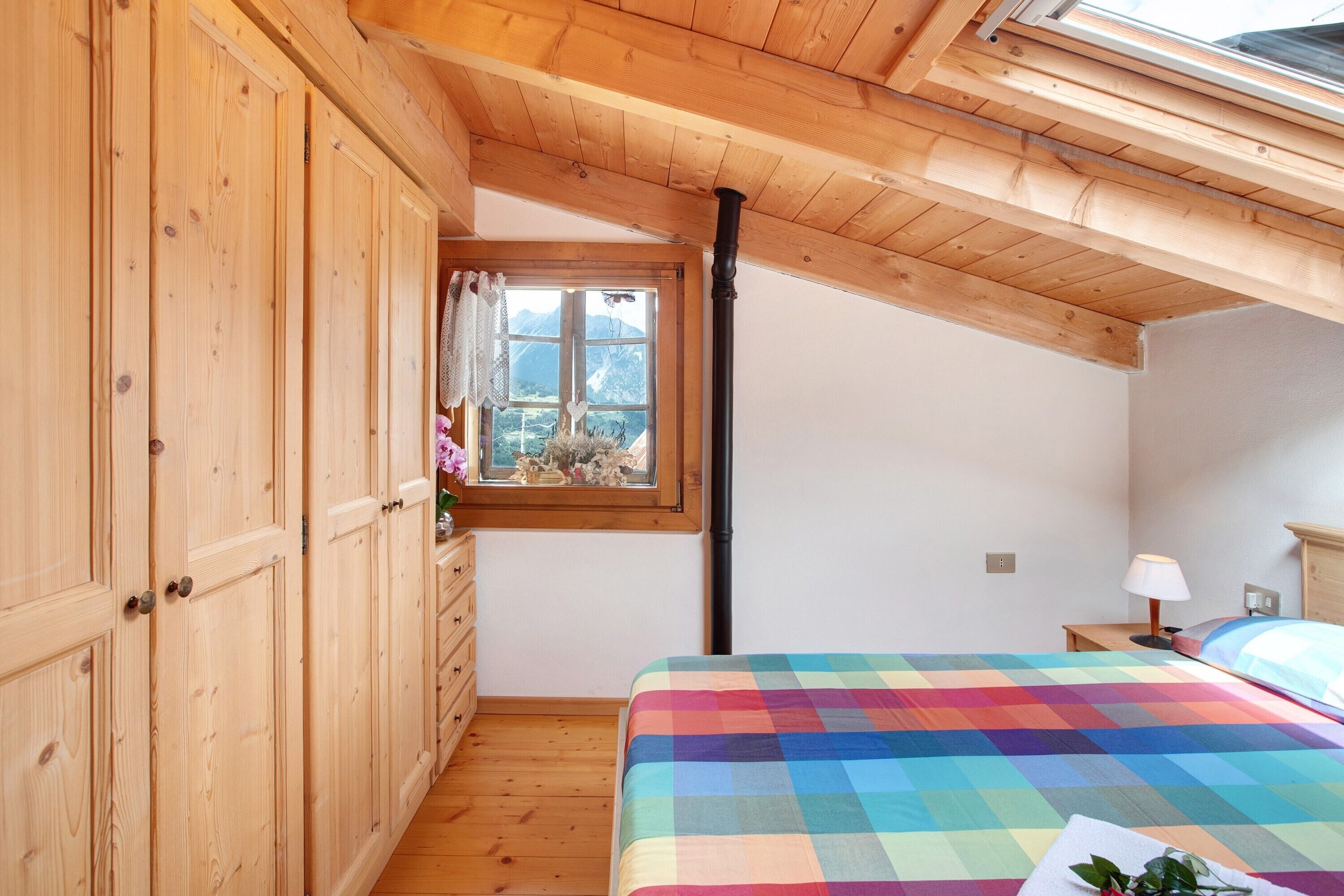 A bedroom with wooden cabinets, a bed with a colorful quilt, a window with a view of mountains, and a skylight.