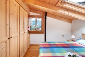 Une chambre avec des armoires en bois, un lit recouvert d’une courtepointe aux couleurs vives, une fenêtre donnant sur les montagnes et un puits de lumière.