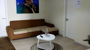 Una sala de estar con un sofá marrón, una mesa de centro blanca con un modelo de barco, una lámpara colgante, un cuadro en la pared que representa una escena costera y una puerta con un letrero de Wi-Fi.