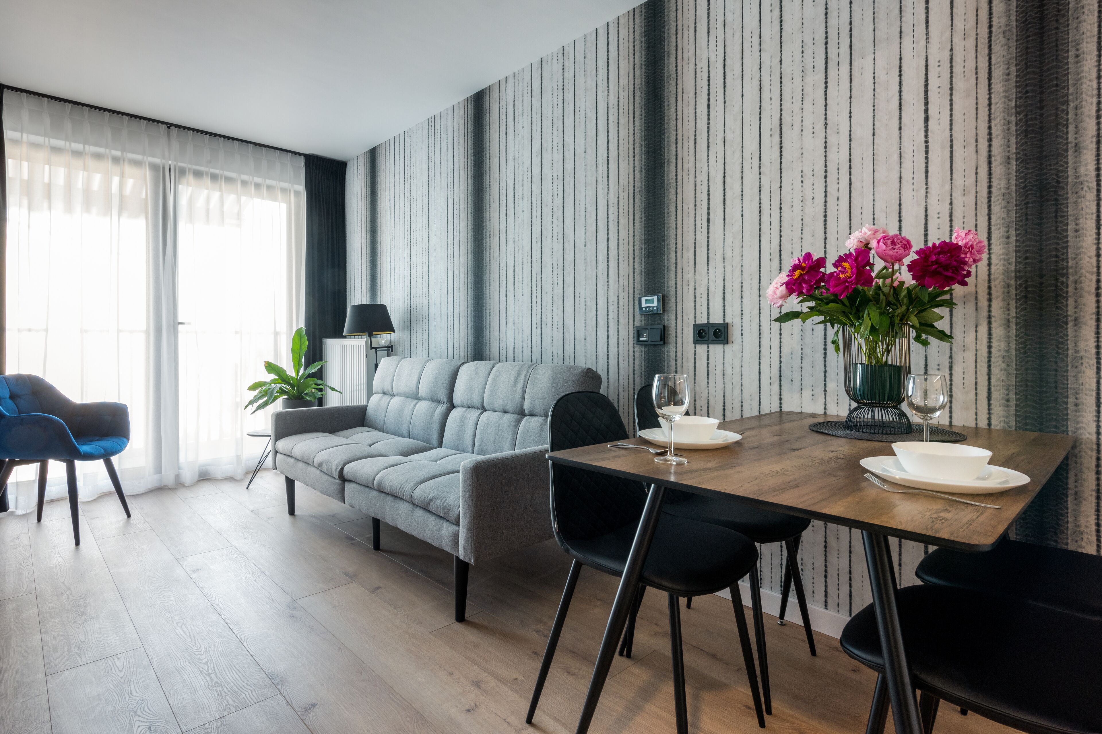 Una sala moderna con un sofá gris, una mesa de comedor con sillas, una planta en maceta y un florero con flores.