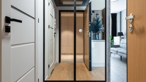 A modern hotel hallway with a wooden floor, a glass door leading to a room, and a view of a living area with a sofa and a lamp. - InPoint Cracow - Serviced Apartments G15 (Kraków)