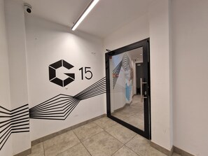 A hallway with a wall-mounted sign displaying a logo and the number 15, a glass door with a geometric design, and a tiled floor. - InPoint Cracow - Serviced Apartments G15 (Kraków)
