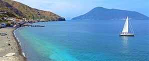 A coastal scene with a clear beach, a sailboat, and a mountain in the background.