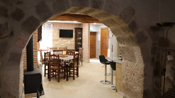 A stone archway leading to a dining area with wooden chairs and a table, a bar area with a stool, and a television mounted on the wall.