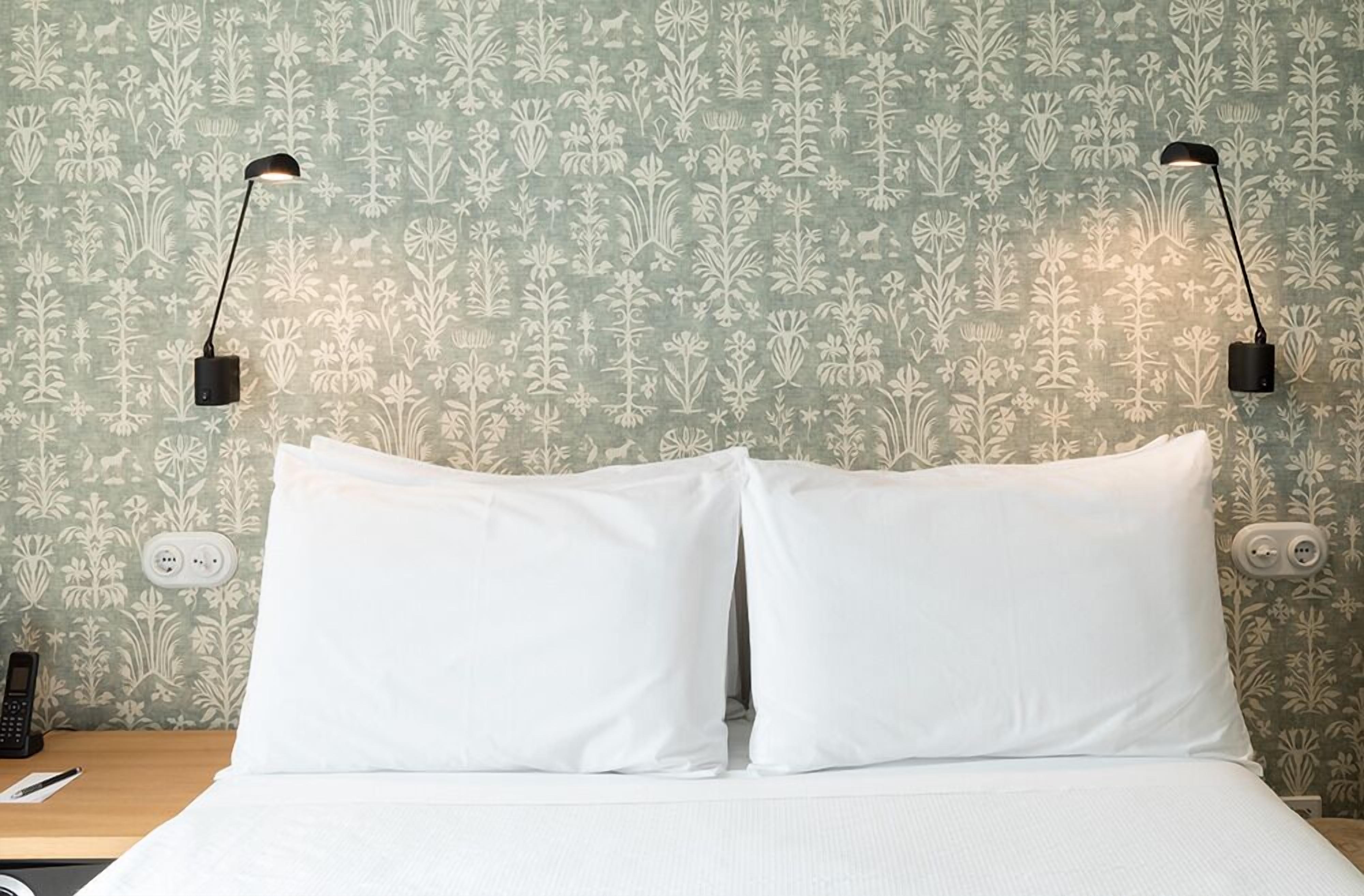 A neatly made bed with white linens, two bedside lamps, and a floral wallpaper pattern.